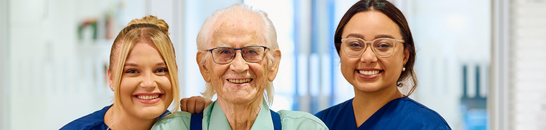 Zwei Pflegerinnen, diese rechts und links von einem älteren Herren stehen. Sie lächeln alle in die Kamera.