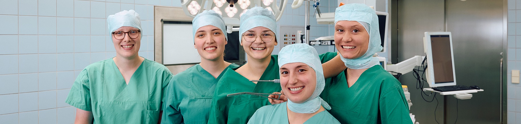 5 junge Frauen, diese in OP Kleidung im OP stehen und in die Kamera lächeln