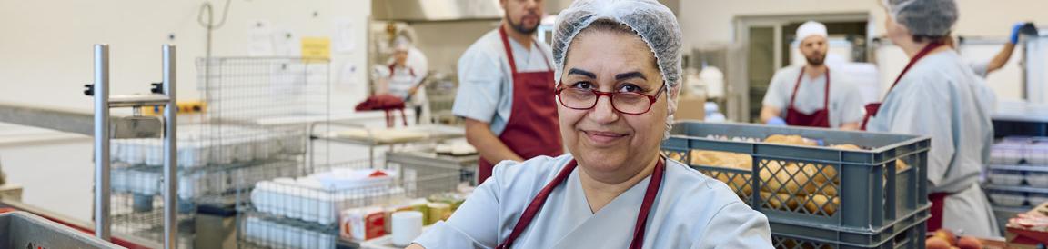 Eine Frau mit Brille, die eine Schutzhaube auf dem Kopf und eine Schürze trägt. Im Hintergrund arbeiten weitere Personen in der Küche